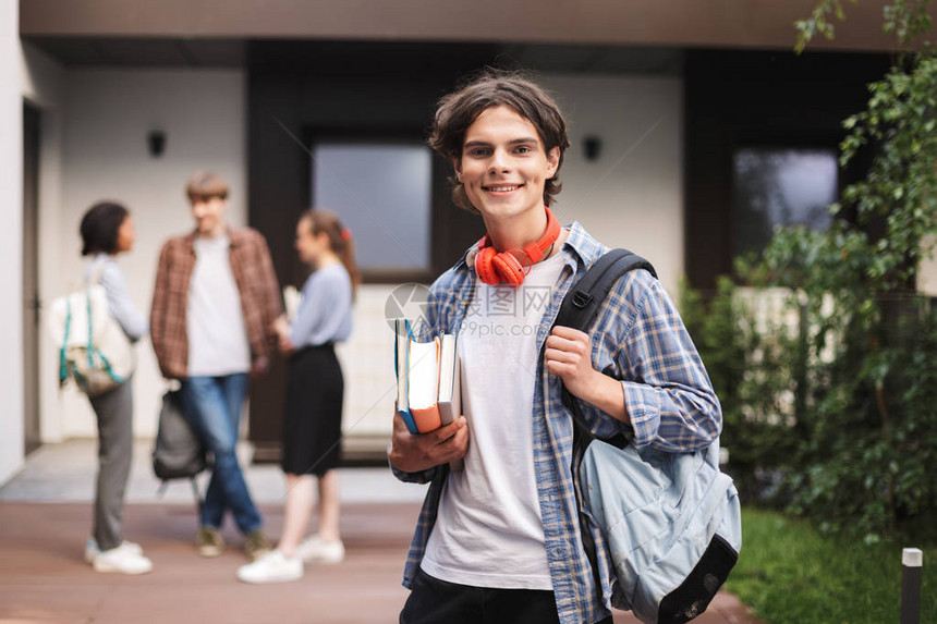 带着红色耳机的快乐男孩背着包和书站在大学的院子里图片