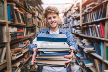 微笑的年轻人在图书馆的背景下手里拿着许多书专注于书籍一名图片