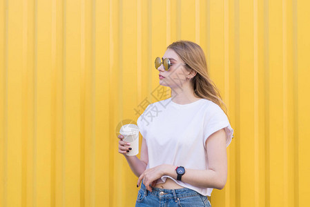 漂亮的女孩在黄色的墙壁背景上手拿一杯咖啡摆姿势在黄色背景上拿着纸杯图片