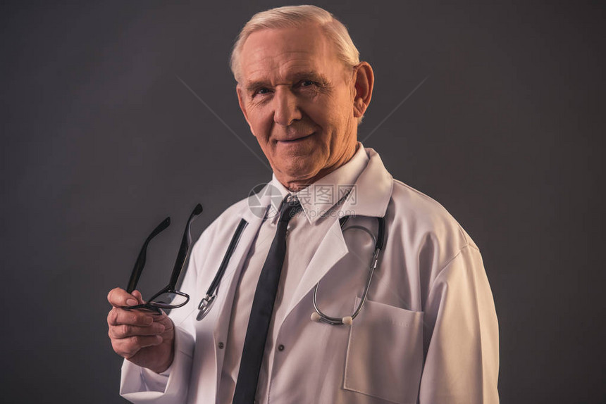 身穿医疗大衣的英俊老医生拿着眼镜用灰色图片