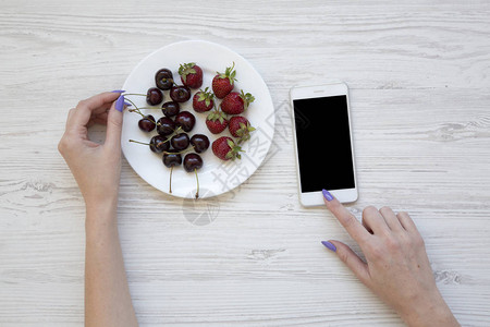 女用智能手机在白木底边吃樱桃和草莓时使用手图片