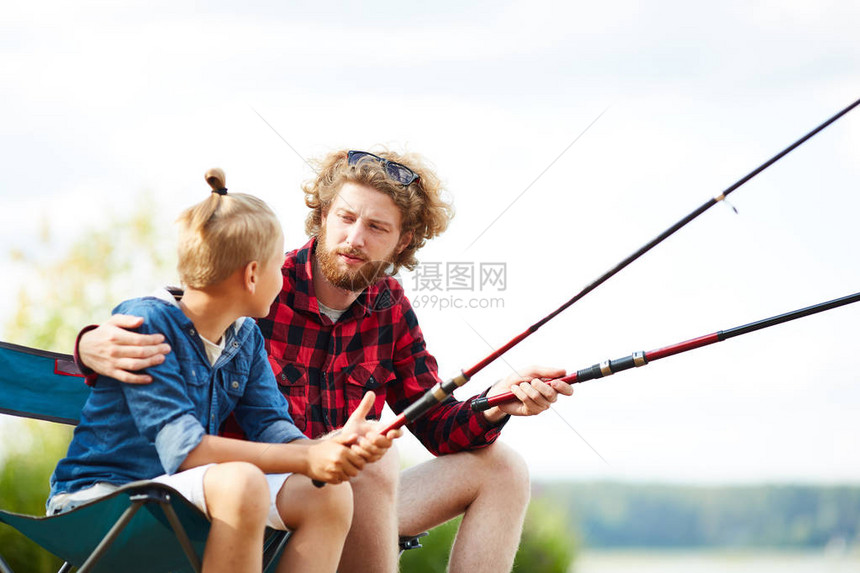 年轻人和他儿子带着钓鱼棒坐在湖边在暑图片