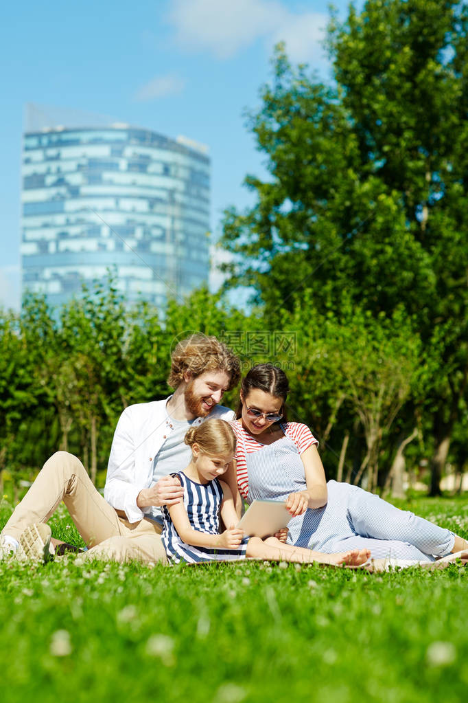 带着触摸板的小女孩和她的父母坐在公园绿草坪上图片