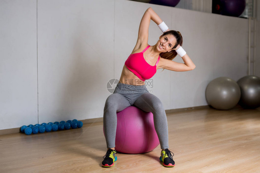 年轻女运动员坐在健身球上用双手在头后图片