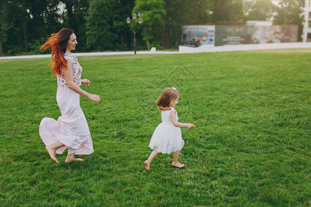 笑着的女人穿着轻便的衣服在公园的绿草坪上追赶可爱的小宝贝女孩图片