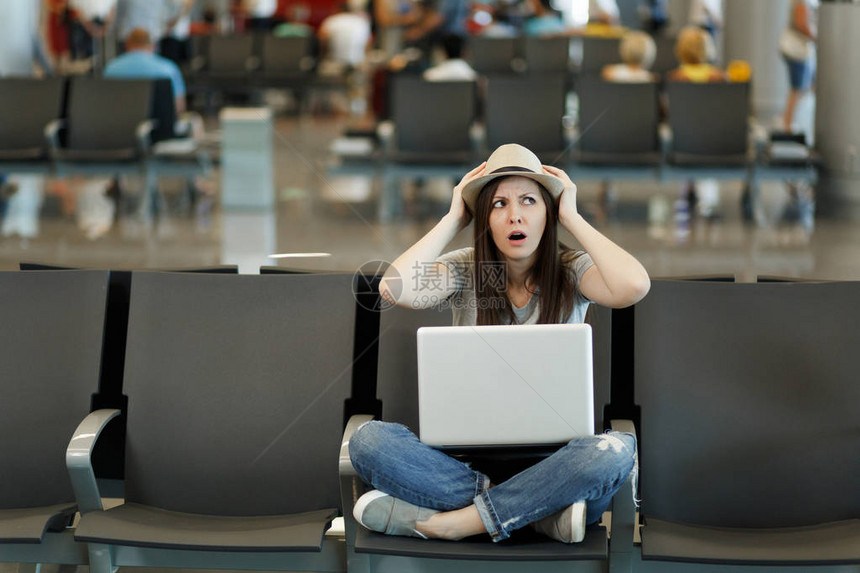 年轻的旅行游女带着笔记本电脑坐在机场大厅等候时盘腿紧握头部图片