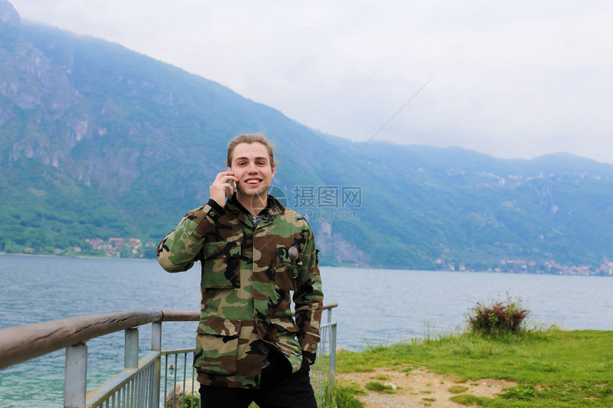 身穿迷彩夹克的白人男游客在栏杆科莫湖和阿尔卑斯山附近用智能手机说话旅游概图片