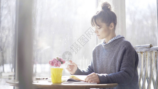 一个穿着蓝色套头衫的黑发严肃女孩正在窗边咖啡馆里的笔图片