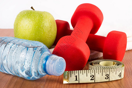 在木制地板上贴近红色哑铃瓶装水绿色新鲜苹果和露珠以及测量胶带运动饮食及健康食背景图片