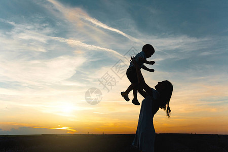 年轻女子的剪影在大自然日落地平线背景上高抛起可爱的小男孩背景