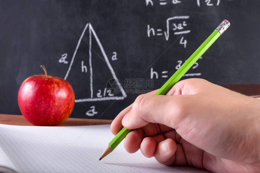 男学生手用绿色木笔在记本上写红苹果和黑板在背景中模糊课堂和教育理念特写图片