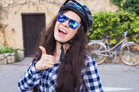 带着头盔和太阳镜的年轻微笑的女人在背景巷子和自行图片