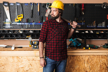 在黄色硬帽的建筑工肩上握着锤子仔细地看着一边拿着工具站住在图片
