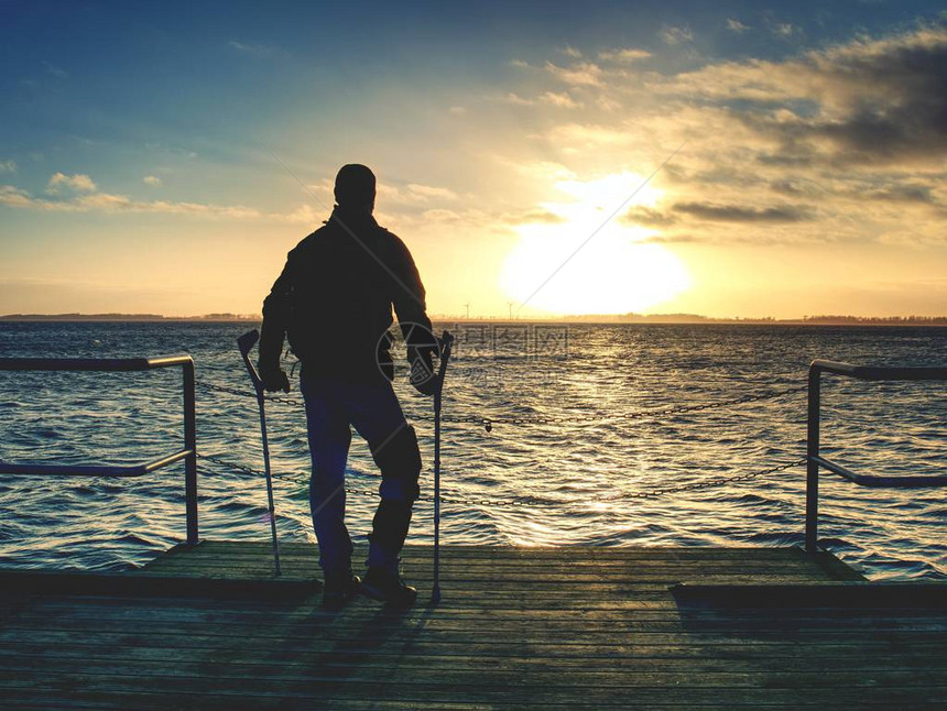 残疾受伤的徒步旅行者正在庆祝他的目标的单独依恋游客在温暖的日落天空接触海水的地平线上站在海桥上或图片