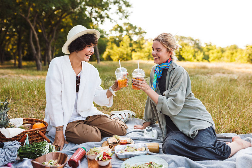 坐在野餐毯子上的两个笑的漂亮女孩把橙汁握在手里快乐地图片
