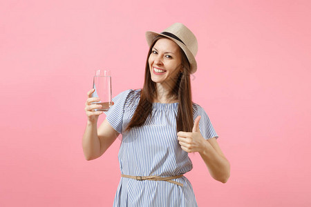 穿着蓝裙子戴帽子和从粉红色背景隔绝的玻璃杯中喝清净纯水的年轻美女肖像背景图片