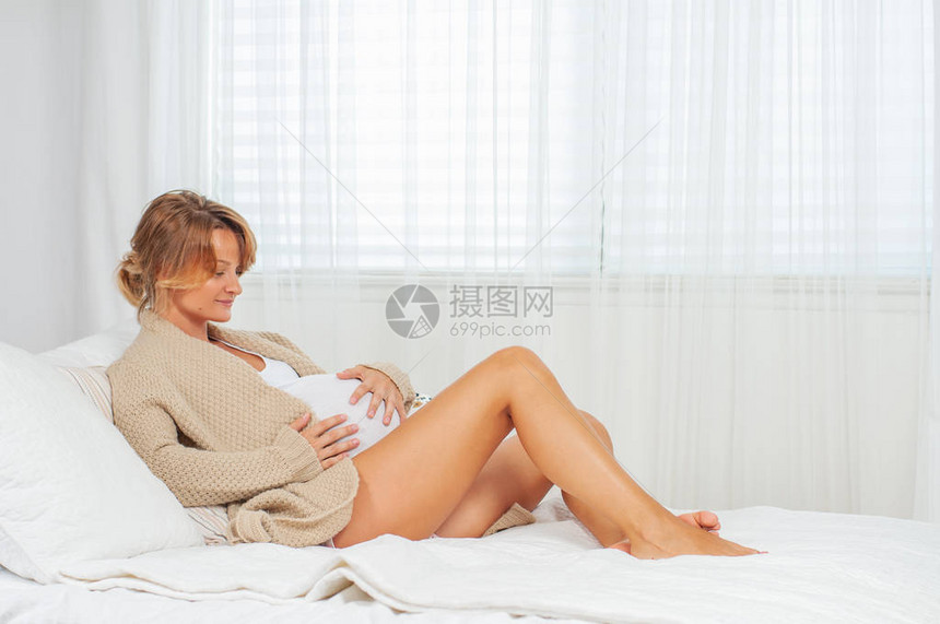 年轻快乐的孕妇用双手抚摸她的肚子就坐在床上图片