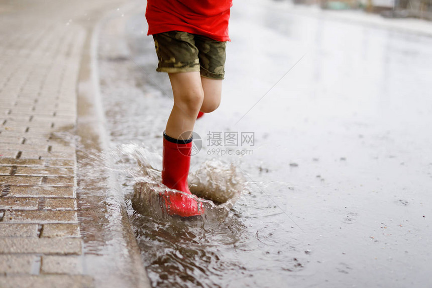穿着红色雨靴的孩子跳进水坑特写孩子玩得开心泼水温暖的夏雨