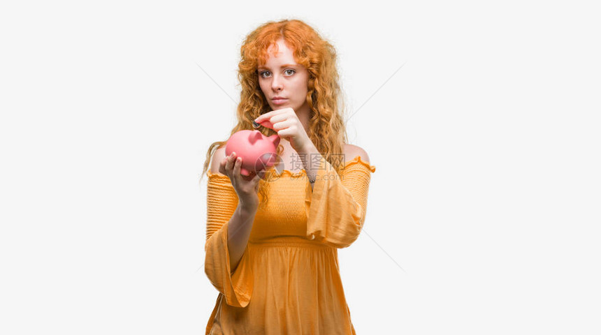 年轻红发女人拿着小猪银行自信地表现在聪明的面孔图片
