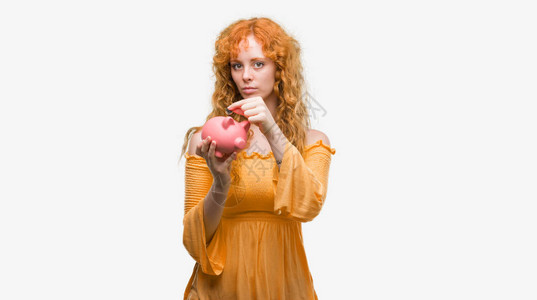年轻红发女人拿着小猪银行自信地表现在聪明的面孔图片