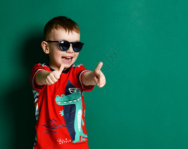 快乐的男孩在太阳镜和红色T恤上展示大拇指标志积极的微笑在浅绿图片