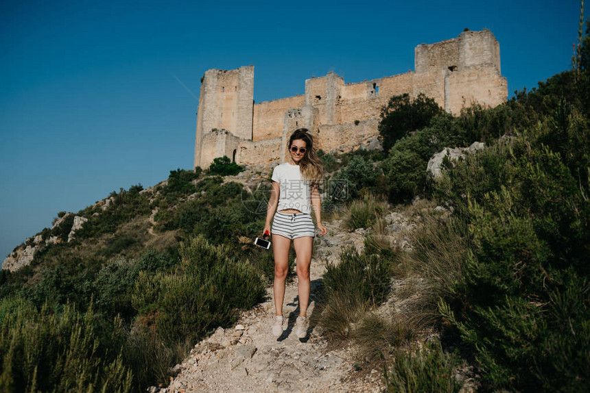 穿着白色t恤和白色短裤的漂亮年轻女子在西班牙日落时分在山上以古城堡为背景在沙路上跳跃图片