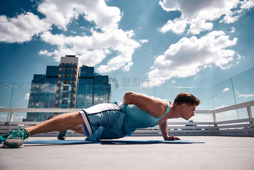 身材匀称的男正在市中心阳光明媚的屋顶上用自己的体重在垫子上锻炼他一边做俯卧撑一边保持平衡男人在露天努力图片