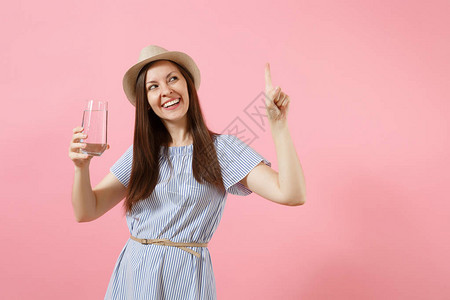 穿着蓝裙子戴帽子和从粉红色背景隔绝的玻璃杯中喝清净纯水的年轻美女肖像背景图片