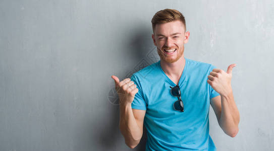 年轻的红发男子在灰色的垃圾墙上穿着休闲装成功的标志用手做积极的手势图片