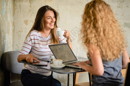 两个女人坐在咖啡馆里边喝咖啡边聊天在桌上的咖啡杯笔记本电脑图片