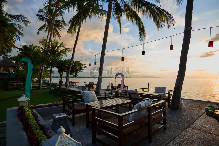 在海岸线的日落热带岛屿岸边的浪漫日落海滩上的餐厅开放式露台餐厅棕榈树下的空桌和椅子背景图片