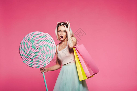 快乐的金发女子拿着巨大的甜棒糖和购物袋装成粉红色背图片