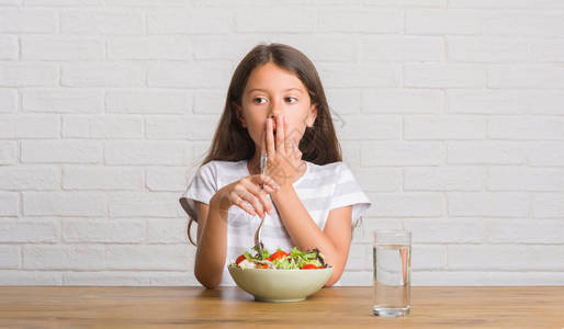 年轻的西班牙裔孩子坐在桌子上吃着健康的沙拉盖着嘴图片