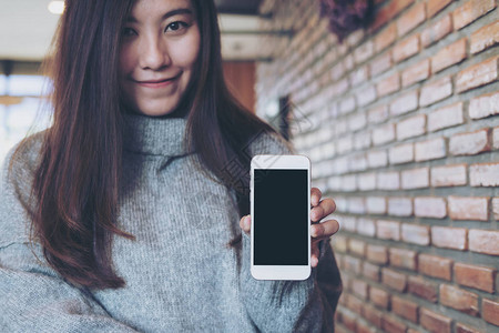 一位亚洲美女在有砖墙背景的咖啡馆中拿着并展示白色手机空白黑屏白黑屏幕的假背景图片
