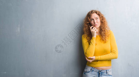 年轻的红发女人在灰色的垃圾墙上用手托着下巴思考问题图片