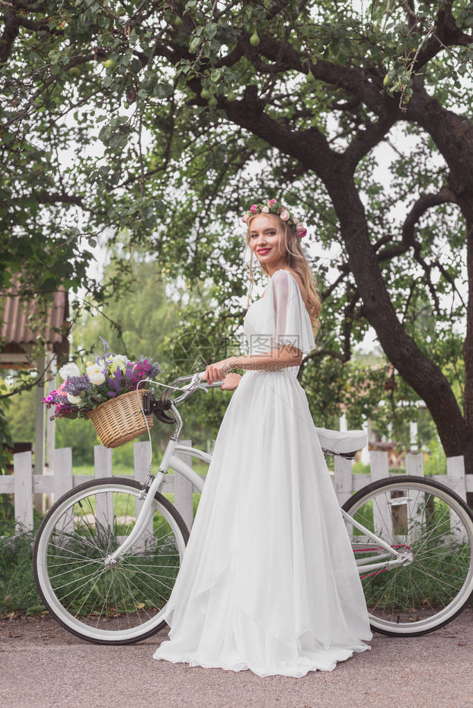 穿着鲜花圈和婚纱的美丽新娘站着脚踏自行车图片