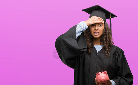 身着毕业制服的年轻西班牙妇女拿着小猪银行头顶着手压力图片