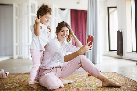 快乐的妈和可爱的小女儿愉快地在手机上拍照在家地毯上共度图片