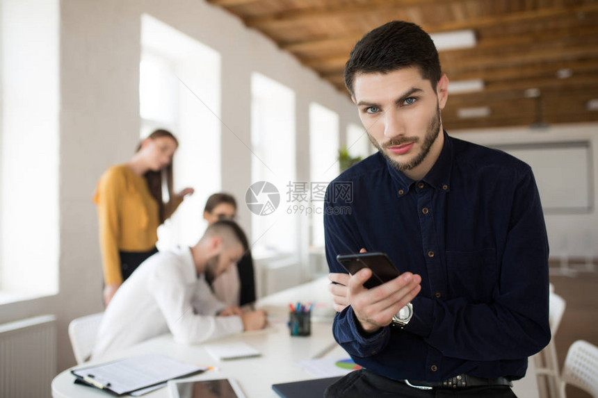 深色衬衫上留胡子的年轻有思想的商人与背景同事在办公室用手机对着办公室进行监视图片