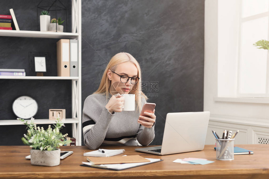 在电脑附近使用智能手机和在办公室复印空间喝咖啡的年轻图片