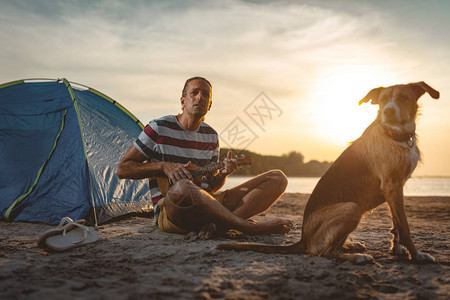 音乐家坐在河边和狗坐在一起在日落图片