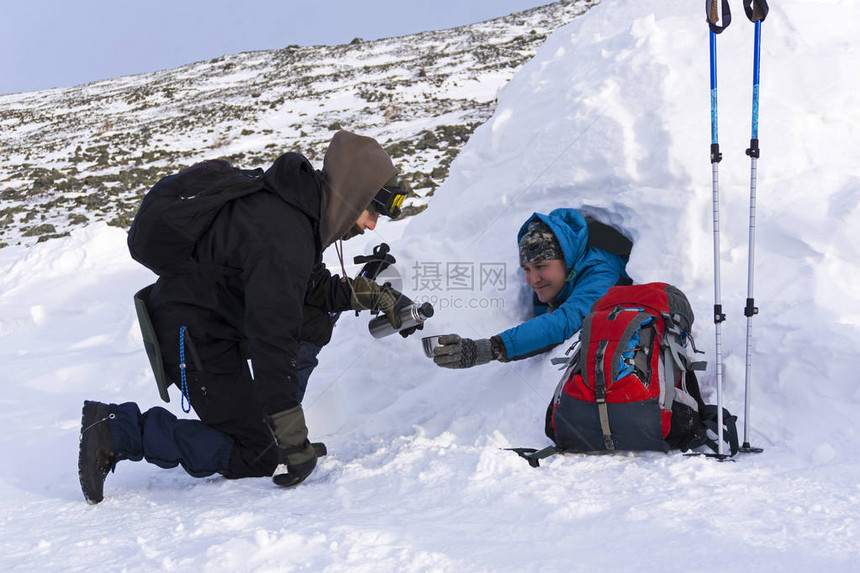 旅行者从温热茶倒到他的朋友在冬季山地风景的背景下坐在雪屋图片