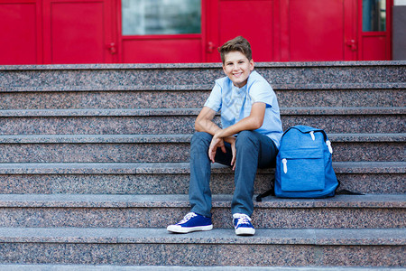 穿蓝衬衫的年轻男孩和蓝色背包坐在学校前面图片