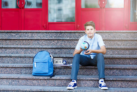 穿蓝衬衫的年轻男孩和蓝色背包坐在学校前面图片