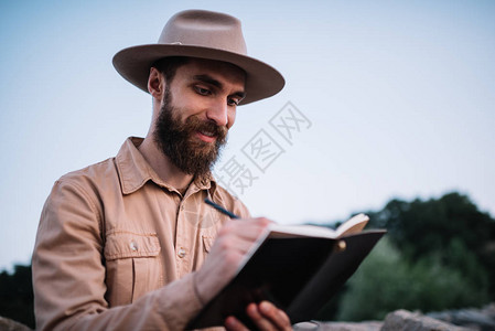微笑的大胡子男戴着时髦的帽子在记事本上写笔记图片