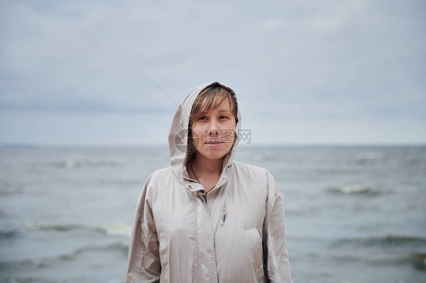 穿着温暖夹克的迷人年轻女微笑着看摄影机站在狂风大海背景上图片