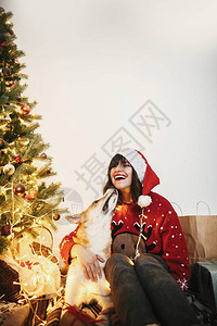 可爱的狗在金色美丽的圣诞树下亲吻戴着圣诞帽的快乐女孩图片