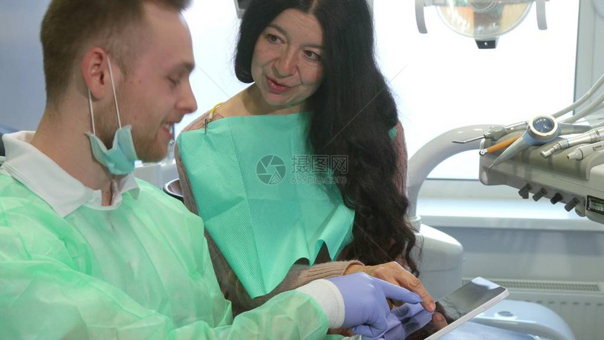 有吸引力的年轻牙医向客户解释他的平板电脑上的东西穿着医疗制服的白种金发男子向他的女病人点头沿牙科保健员和高级图片