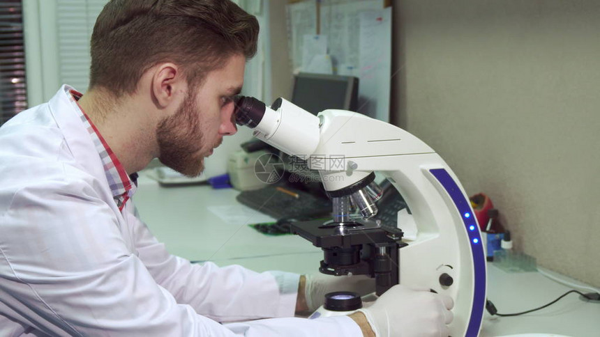 有魅力的年轻人穿着白大褂透过实验室的范围看白种人男科学家通过显微镜探测一些测试材料的侧视图黑发有胡子的实图片