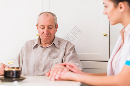 坐在厨房桌子旁的老病人和坐在他旁边握着他的图片
